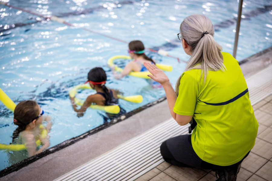 Teacher on the side of the pool giving instructions to three young swimmers with pool noodles in the water