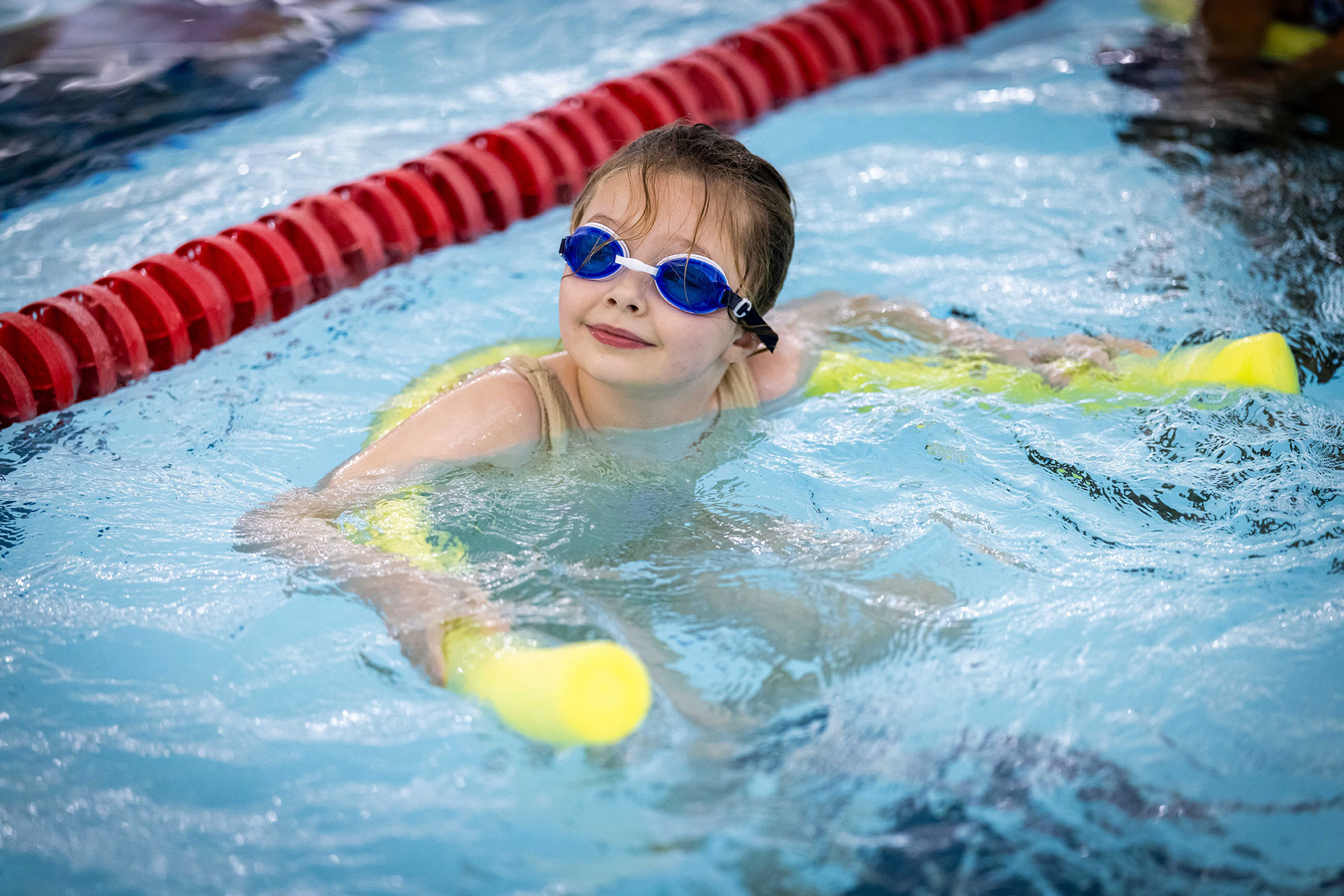 A young girl in the swimming pool wearing goggles and using a floatation aid.