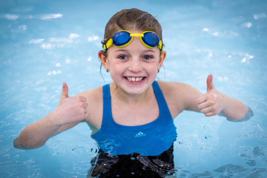 young grile in a swimming pool with goggles on her head giving two thumbs up